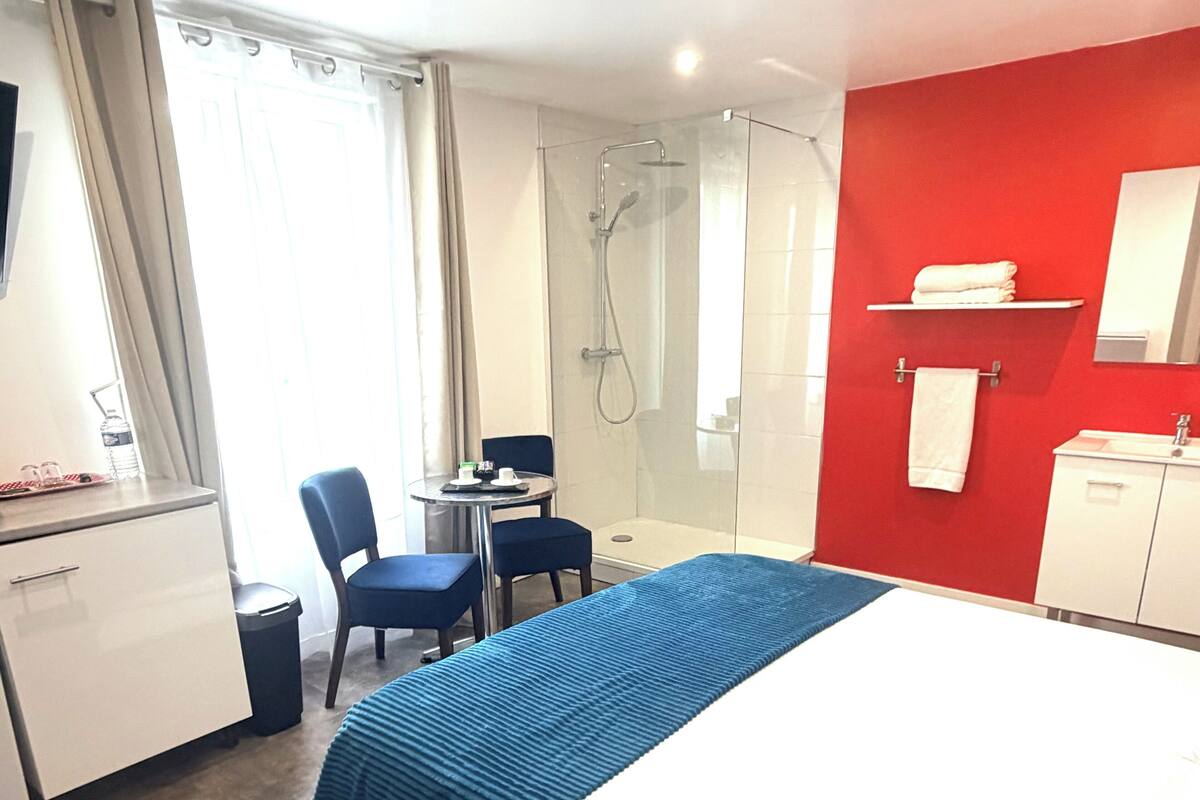 A hotel room with a bed, a desk with chairs, a shower, a sink, and a red accent wall.