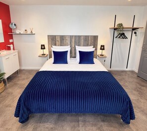 A bedroom with a large bed, two bedside tables, a red accent wall, a wooden headboard, and a hanging plant. -  Aux Charmes (Isques)