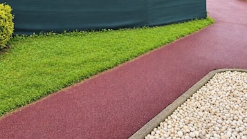Un chemin recouvert d’un revêtement rouge, bordé d’une haie verte et d’une zone de gravier.