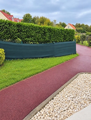 A pathway with a red surface, bordered by a green hedge and a gravel area. -  Aux Charmes (Isques)
