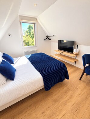 A bedroom with a bed, a TV, a chair, and a window with a view of trees. -  Aux Charmes (Isques)