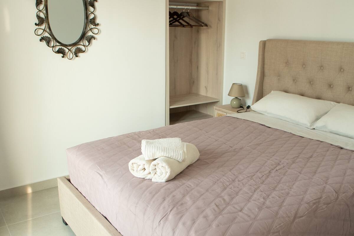 A neatly made bed with a quilted cover, a bedside table with a lamp, a wooden wardrobe, and a wall-mounted mirror.