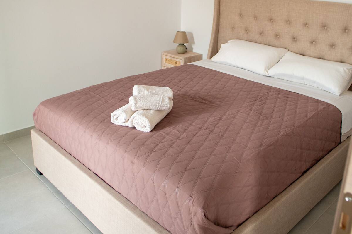 A neatly made bed with a pink quilt, a wooden headboard, and a bedside table with a lamp.
