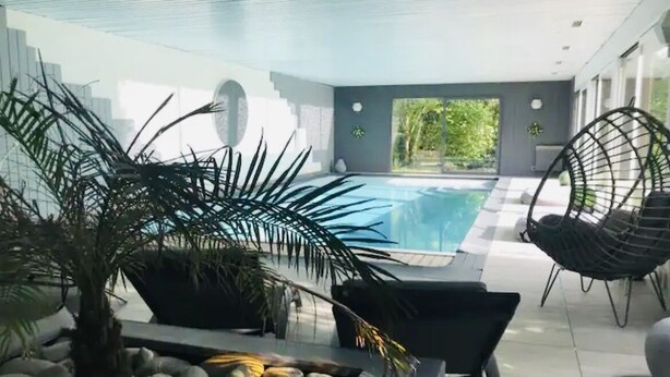 A modern indoor pool area with a clear view of the pool, surrounded by a minimalist design, including a palm tree, a black chair, and a seating area with a table. - Magnificent residence with heated indoor pool (Guipry-Messac)