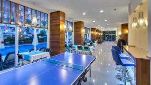 A modern indoor dining area with tables and chairs, a ping pong table, and large windows with blinds. - Novella Apart Hotel (Alanya)