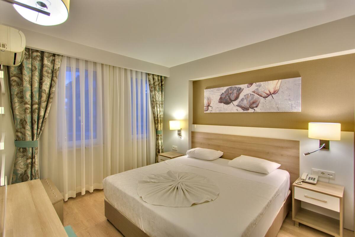 Ein Hotelzimmer mit einem großen Bett, einem Nachttisch mit Lampe, einem weiteren Nachttisch mit Telefon und einem Fenster mit Vorhängen.