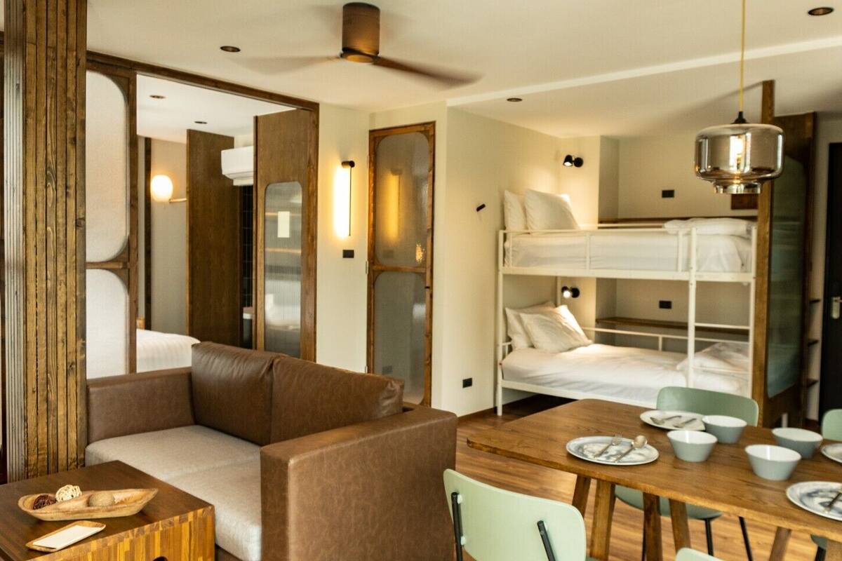 A compact hotel room with a bunk bed, a sofa, a dining table with chairs, and a ceiling fan.