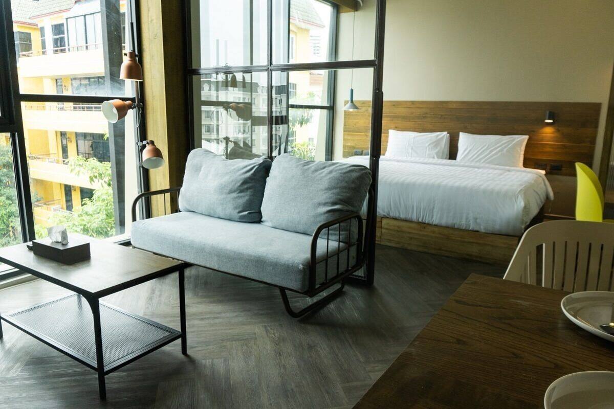 A modern hotel room with a grey sofa, a wooden bed, a small table, and a chair.