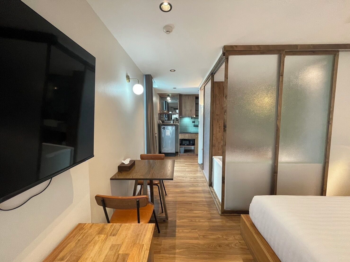 A compact hotel room with a wooden dining table, a single bed, and a kitchenette in the background.
