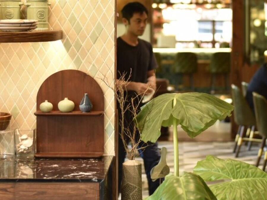 A restaurant interior with wooden tables and chairs, a counter with various dishes, and a large green plant in the center.