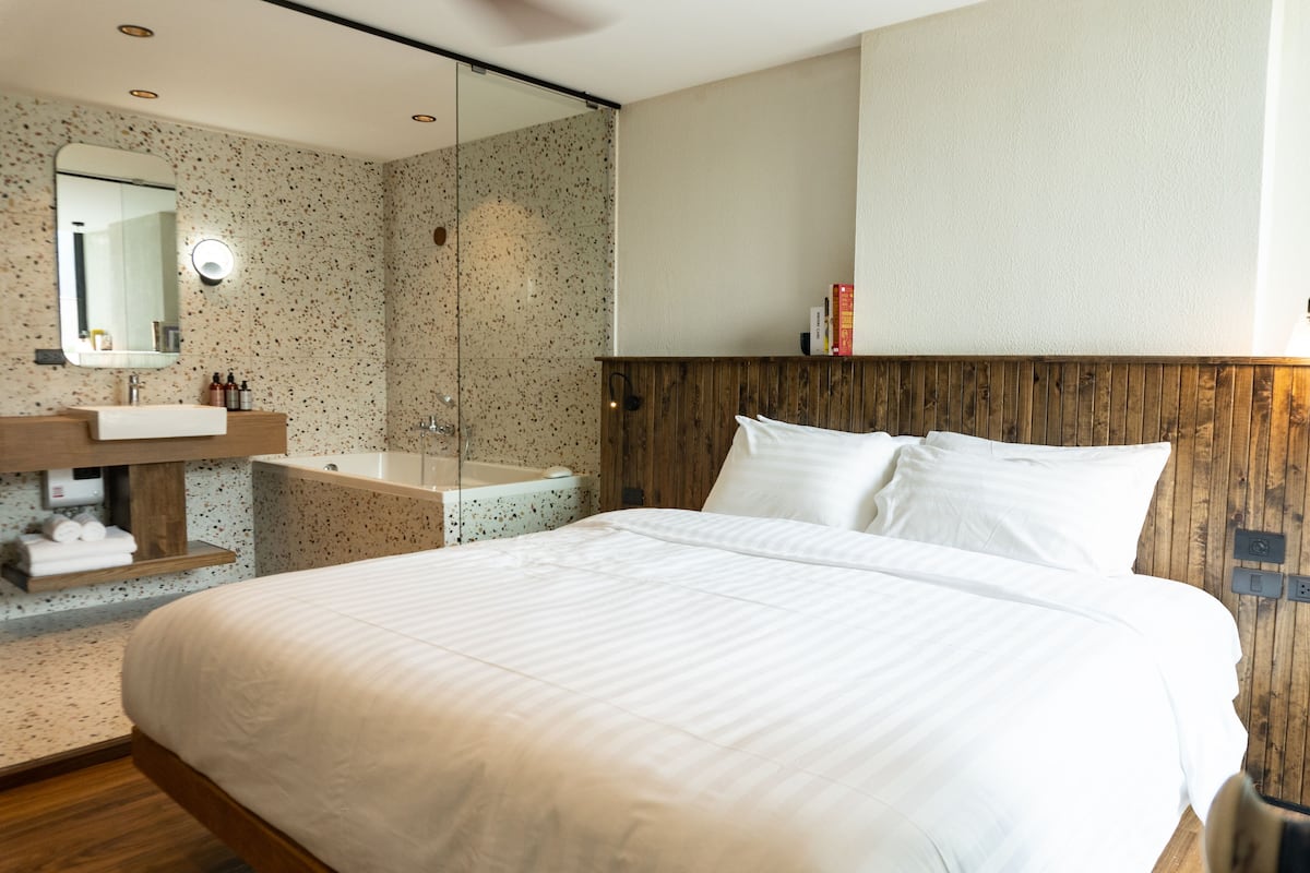 A modern hotel room with a large bed, a wooden headboard, a glass shower enclosure, and a wooden vanity with a mirror.