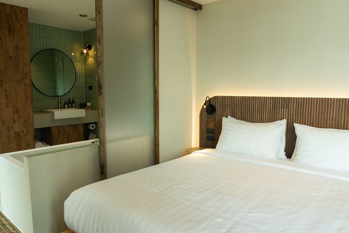 Habitación de hotel con cama, cabecera de madera, espejo redondo y un baño pequeño con lavabo y artículos de aseo.
