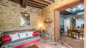 A rustic room with a stone wall, a wooden bench, a red sofa, a table with a lamp, and a dining area with a wooden table and chairs. - Amazing home in Castiglion Fiorentino (Castiglion Fiorentino)