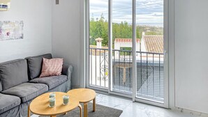 En moderne stue med en grå sofa, to træsideborde og en stor skydedør, der fører ud til en balkon med udsigt til bygninger og træer.