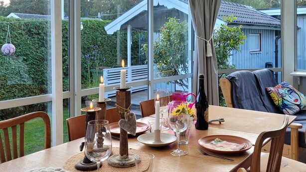 A dining table set with candles, glasses, and a bottle, overlooking a garden and a house.