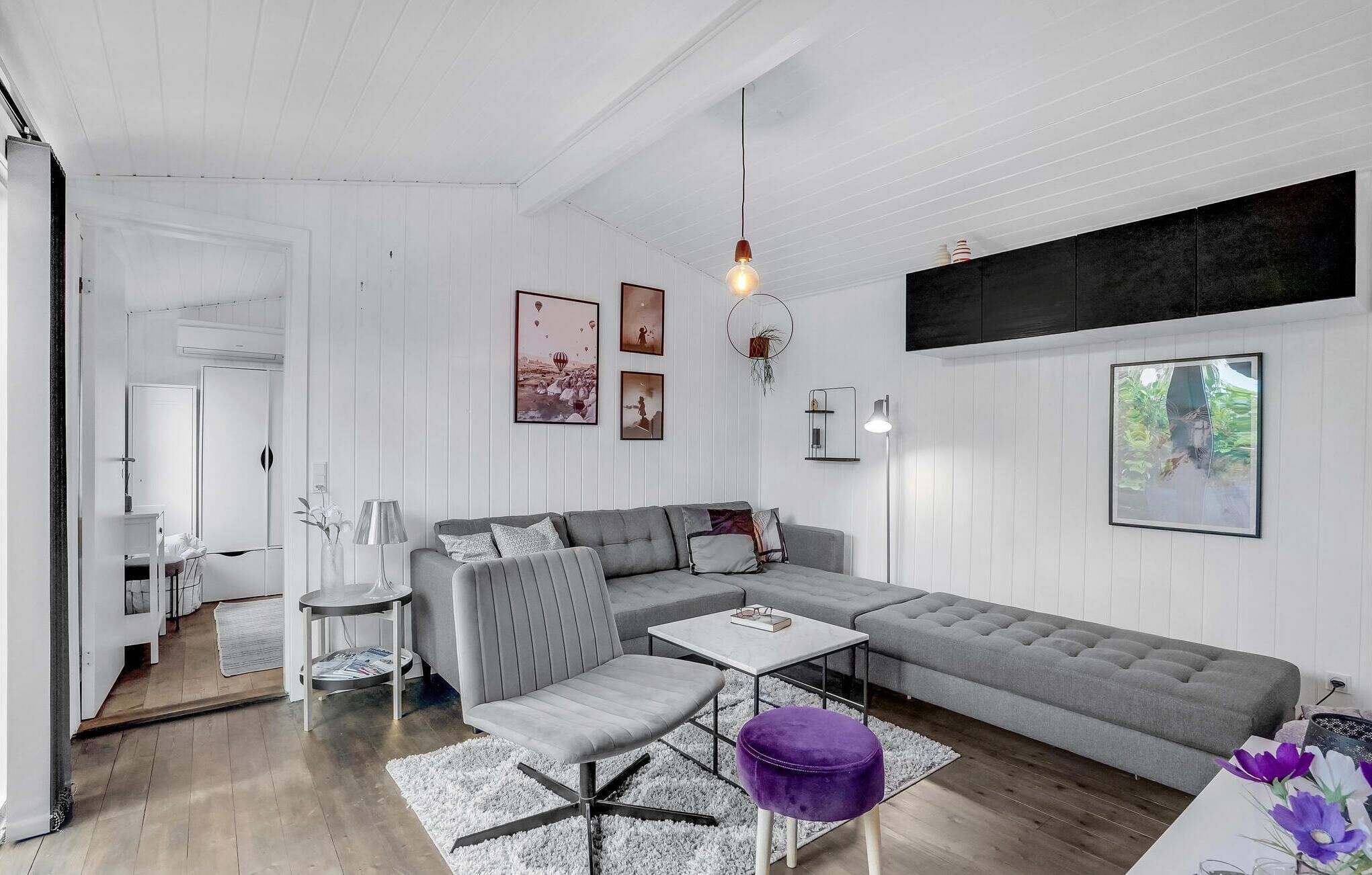 Una sala moderna con un sofá seccional gris, un sillón gris, una pequeña mesa de centro blanca y un taburete púrpura. Hay un mueble negro grande sobre el sofá y un cuadro en la pared.