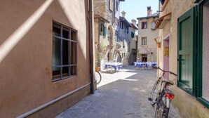 A narrow cobblestone street with a bicycle parked by a building with green shutters. Tables and chairs are set up outside a restaurant. - 2 bedroom cozy apartment in Grado (Grado)