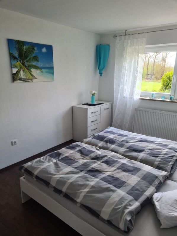 Un dormitorio con cama, mesita de noche, ventana con cortinas y un cuadro en la pared.