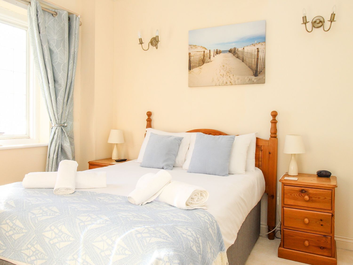A bedroom with a bed, two bedside tables, a window with curtains, a wall-mounted picture, and a nightstand with a lamp.