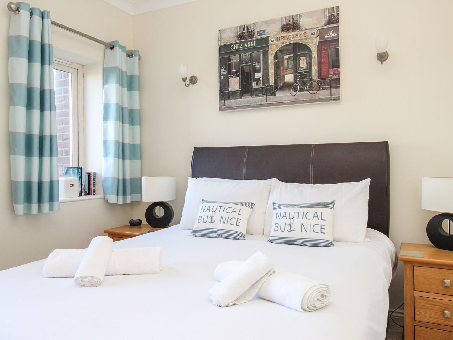 A neatly made bed with two pillows, a window with curtains, a wall-mounted picture, and a nightstand with a lamp.