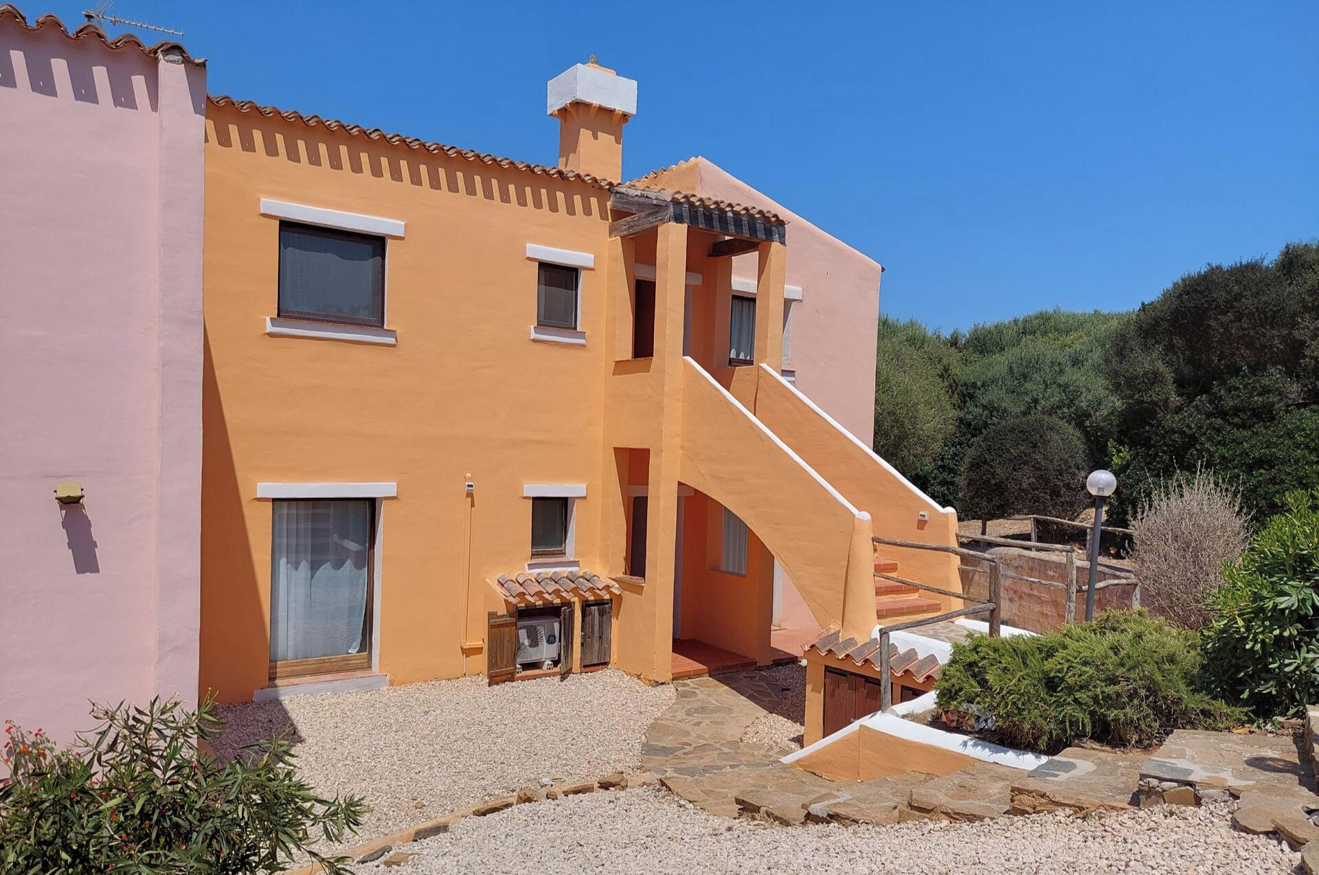 A two-story building with a pink facade and orange accents, featuring a central staircase, a small outdoor seating area, and a gravel driveway.