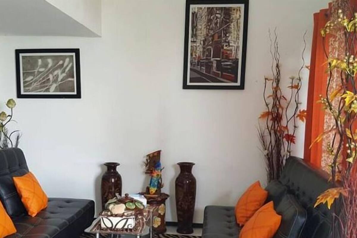 A living room with a black sofa, orange cushions, and a coffee table with decorative items. There are framed pictures on the wall and a large vase with branches.