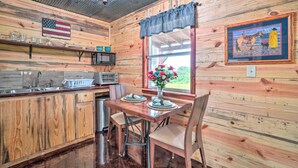 A rustic kitchen with wooden walls, a dining table with chairs, a window with a view, and an American flag on the wall. - Updated Studio Cabin in Ozark w/ Yard & Mtn View (Ozark)
