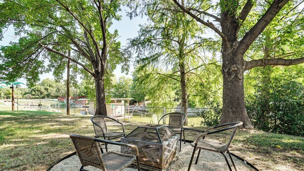 Ein Sitzbereich im Freien mit Korbstühlen und einem Metalltisch, umgeben von Bäumen und einem eingezäunten Sportplatz.