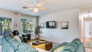 Ein Wohnzimmer mit einem großen, grünen Sofa, einem Couchtisch und einem Deckenventilator. An der Wand hängen zwei gerahmte Bilder, und über einem Holztisch ist ein Fernseher angebracht.