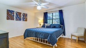 Ein Schlafzimmer mit Bett, einem Sessel, einem Fenster mit Vorhängen, einem Deckenventilator und Bildern an der Wand.