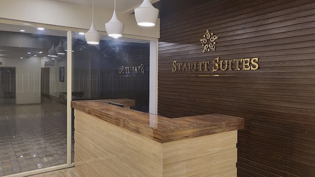 A reception desk with a wooden finish and a glass partition. Above the desk, there are three pendant lights. The wall behind the desk has a dark wood paneling with the text "Starlit Suites" in gold lettering. - STARLIT SUITES SHIRDI (Rahata)