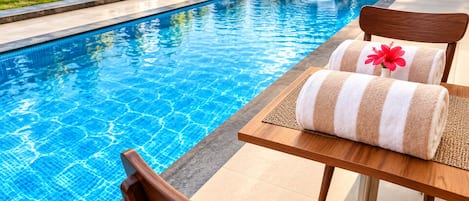 A poolside setting with a wooden table and chairs, a towel with a flower, and a clear blue pool.