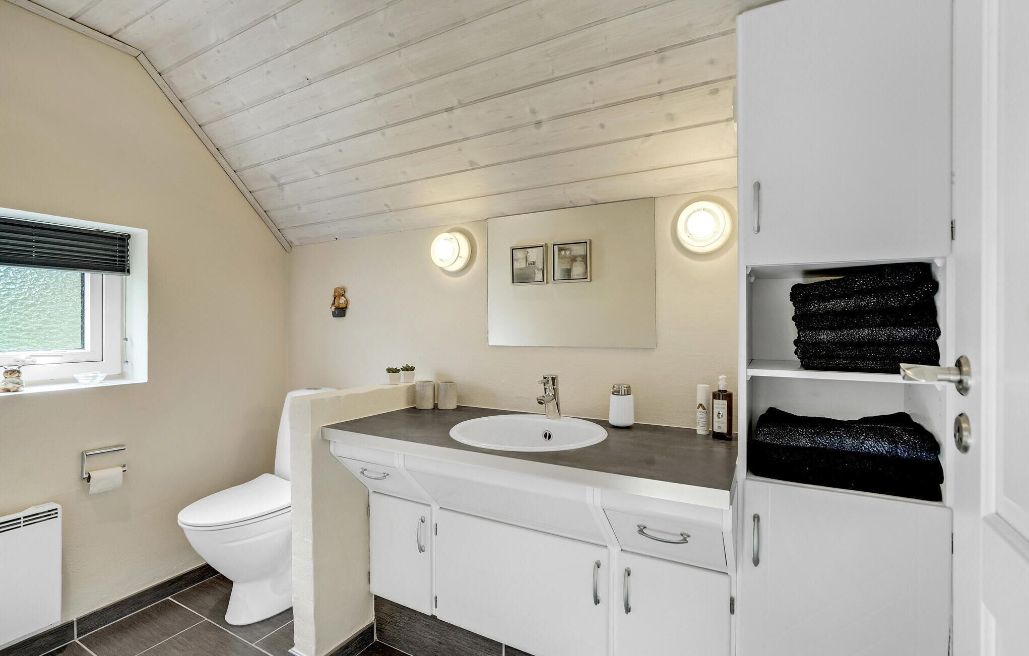 Ein modernes Badezimmer mit einem weißen Waschbecken, einem Waschtisch und einem Spiegel darüber. Eine Toilette befindet sich links, rechts eine Ablagefläche mit gefalteten Handtüchern.