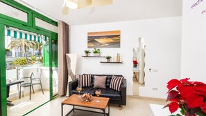 A modern living room with a black leather sofa, a coffee table with wine glasses, a balcony with a view of palm trees, and a painting on the wall.