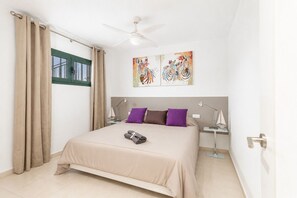 Une chambre à coucher comprenant un lit, deux lampes de chevet, un ventilateur de plafond, une fenêtre avec des rideaux et deux œuvres d’art accrochées au mur.