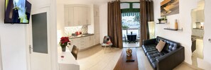 Un salon moderne avec un canapé en cuir noir, un coin repas avec une table et des chaises, une cuisine avec des meubles blancs et un téléviseur fixé au mur.