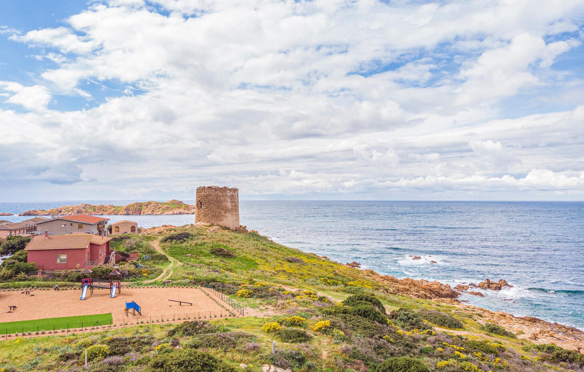 A coastal landscape with a historic tower, a sandy beach with playground equipment, and residential buildings.