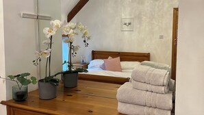 Une chambre à coucher avec une commode en bois, un miroir, des orchidées blanches, un lustre et un lit bien fait, agrémenté d’oreillers roses et blancs.