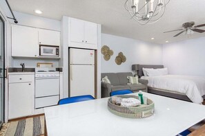 A compact hotel room with a kitchenette, a bed, a seating area, and a ceiling fan.