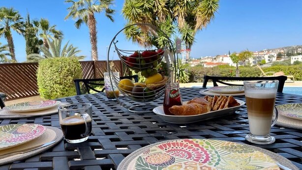 Une table dressée à l’extérieur, avec vue sur des palmiers et des maisons, où l’on aperçoit des assiettes de nourriture, un verre de bière et une bouteille d’eau.