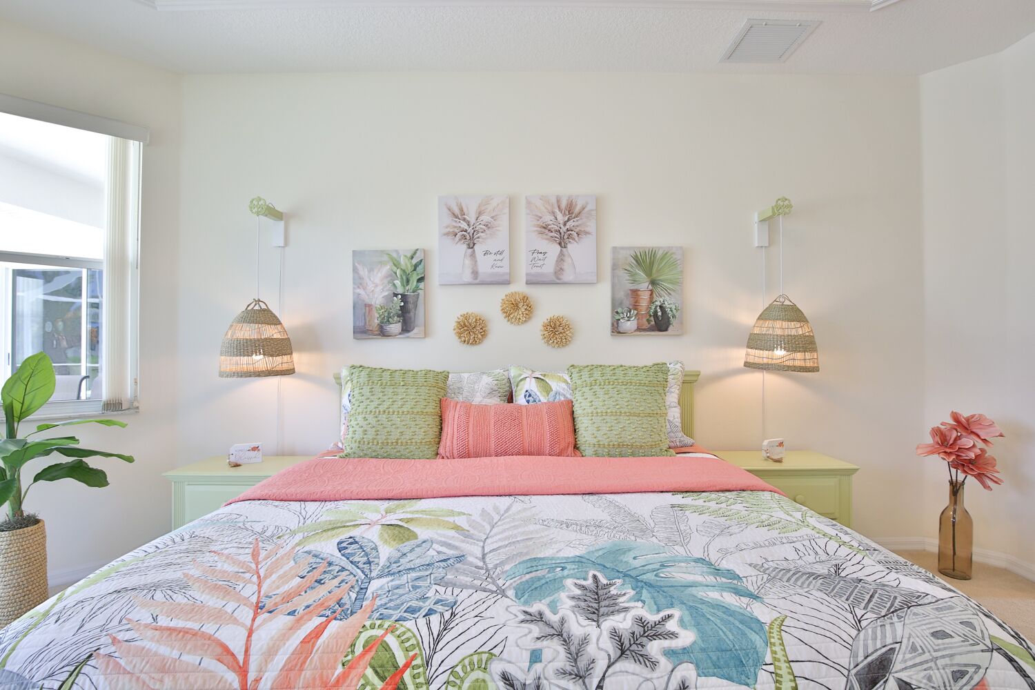 A bedroom with a floral-patterned bedspread, green and pink pillows, and wall-mounted lamps.