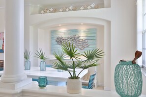 Un intérieur moderne avec une colonne blanche, une œuvre d’art murale aux motifs bleus et blancs, et des plantes en pot disposées sur une table blanche.