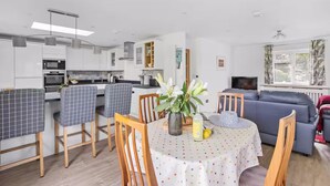 Eine moderne Küche mit weißen Schränken, eine Essecke mit Tisch und Stühlen, eine Sitzecke mit Sofa und ein Fernseher in einem Wohnzimmer.