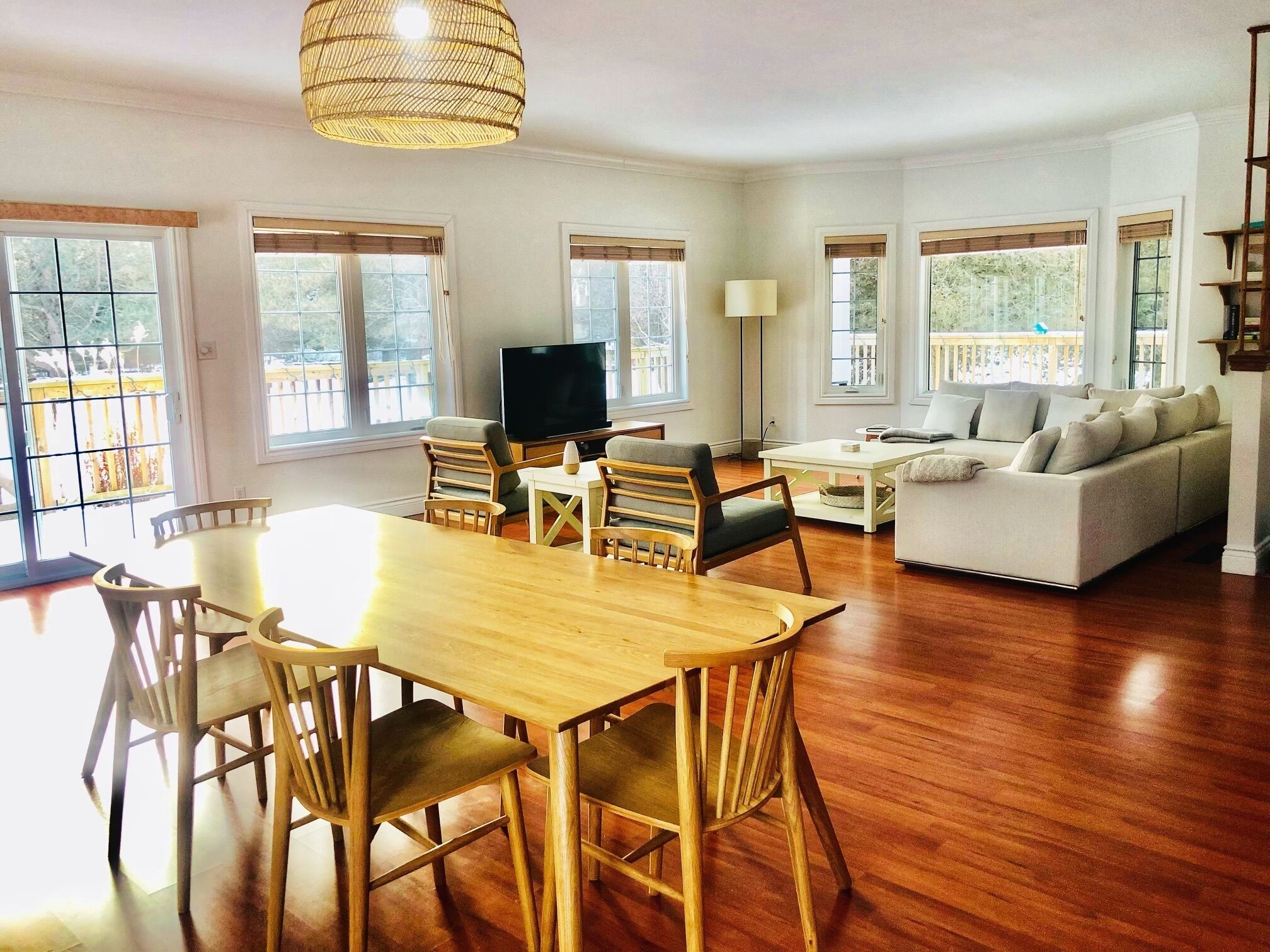 A spacious living room with a wooden dining table, chairs, a sofa, and a TV. Large windows provide natural light and views of the outdoors.
