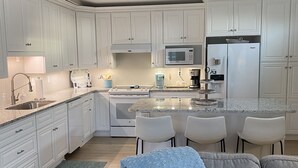 A modern kitchen with white cabinetry, a central island with bar stools, a microwave, and a refrigerator. - 3 BR, 2 Bath home in Emerald Isle! Golf cart, beach chairs & Shibumi included! (Emerald Isle)