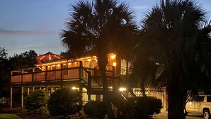A two-story house with a deck and a covered patio area, surrounded by palm trees and a lawn. - 3 BR, 2 Bath home in Emerald Isle! Golf cart, beach chairs & Shibumi included! (Emerald Isle)