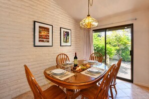 A dining area with a brick wall, a glass table, wooden chairs, a chandelier, and a view of greenery through a glass door.