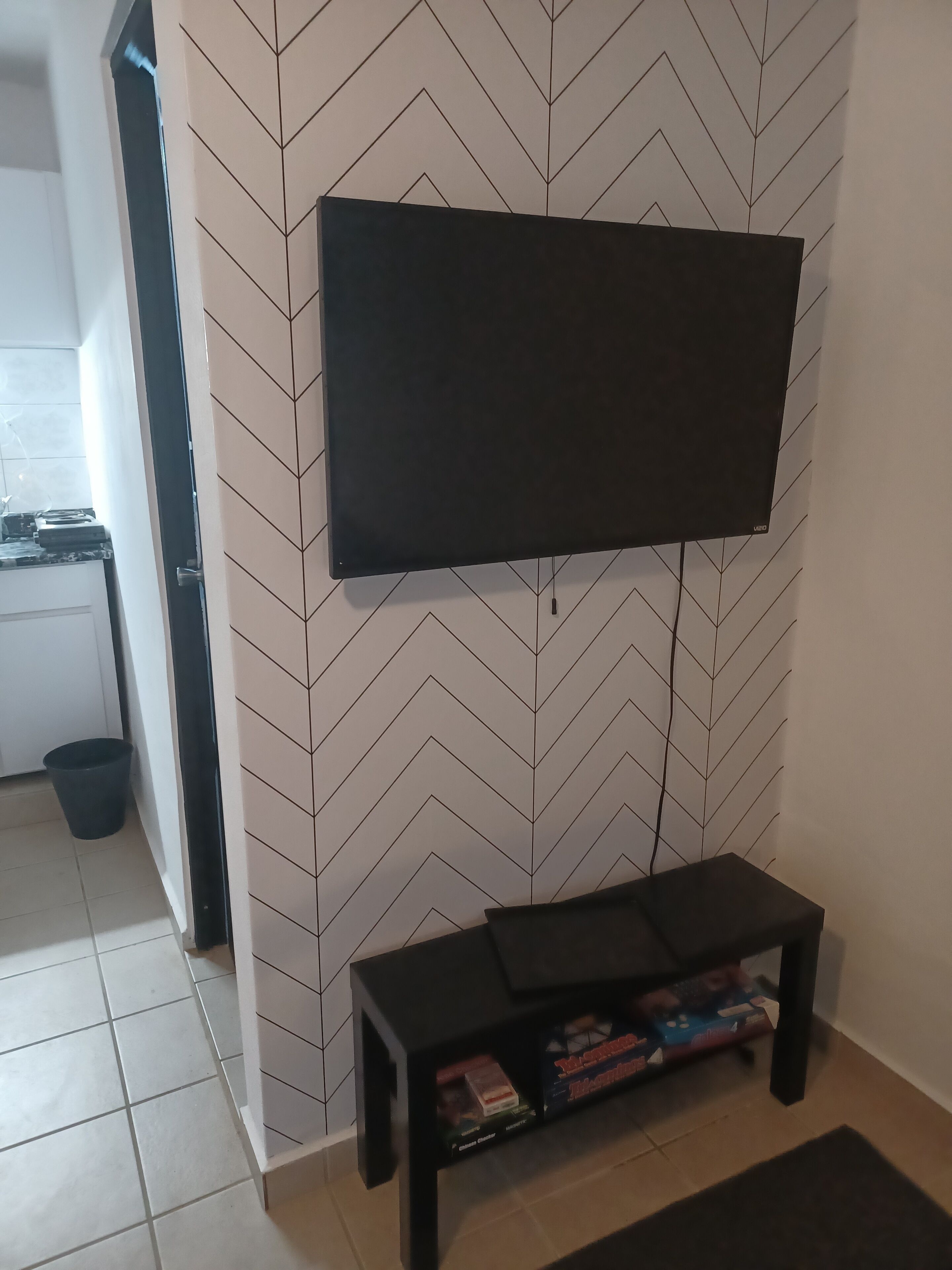 A living room with a flat-screen TV mounted on a chevron-patterned wall, a black entertainment unit with books and games, and a small black bench.