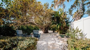 A paved pathway leading to a seating area with two chairs, surrounded by lush greenery and palm trees. - TranquiliSea: Pet friendly, long term discount, resort-style pool! (Holmes Beach)
