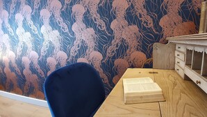 Une pièce avec une chaise bleue, un bureau en bois et une bibliothèque adossée à un mur recouvert d’un papier peint à motifs floraux.